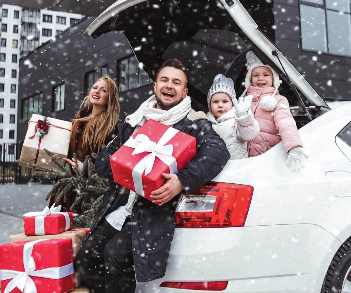 Family of four holding a lot of gifts in snowy weather in front of a car trunk.