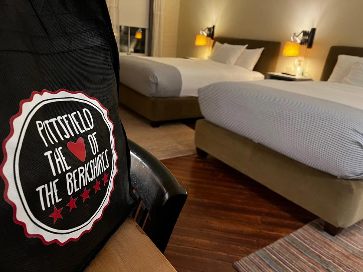 A cloth bag reading "Pittsfield the heart of the Berkshires" sits on a hotel table with hotel beds in the distance.