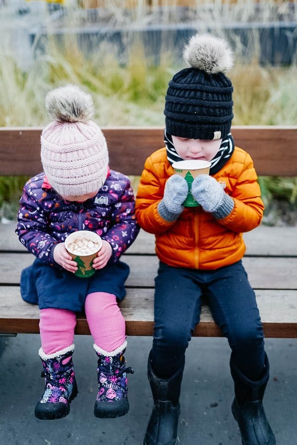 Two kids sip on hot chocolate, while sitting on a bench at the St. Paul European Christmas Market, one of the best christmas markets in the United States for families.