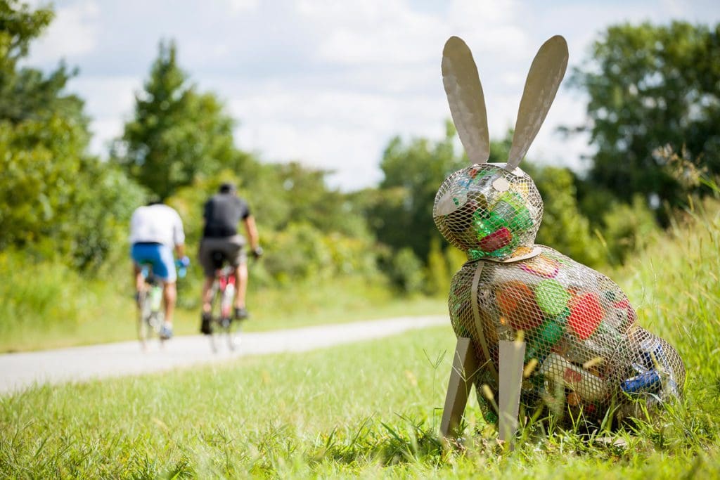 Two people bike along the Swamp Rabbit Trail in the background, with a recycled material bunny art installation in the foreground.
