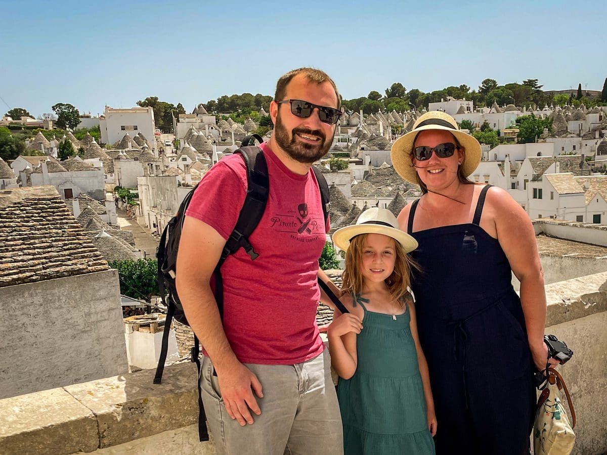 A family of three stands together with the iconic trulli rooftops of Alberobello in the distance.