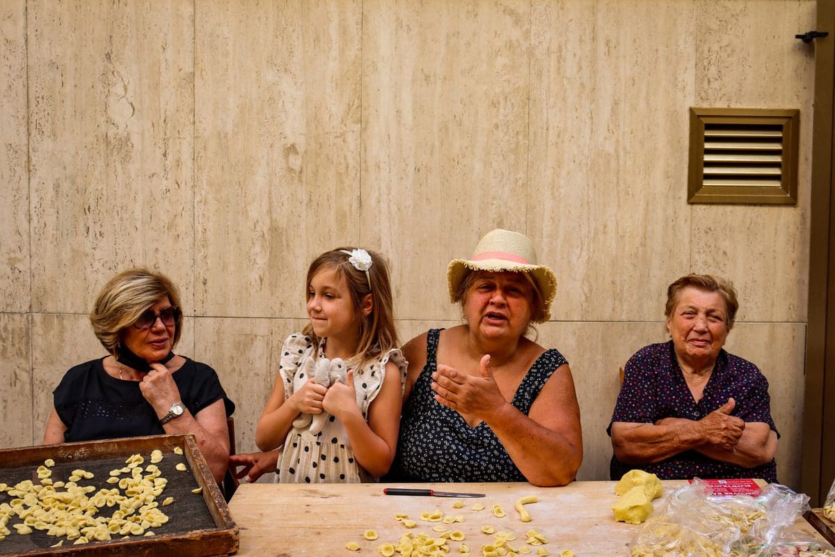 A young girl sits with three Italian women making pasta on a street in Bari, knowing which cities to visit is one of the essential tips for visiting Puglia with kids.