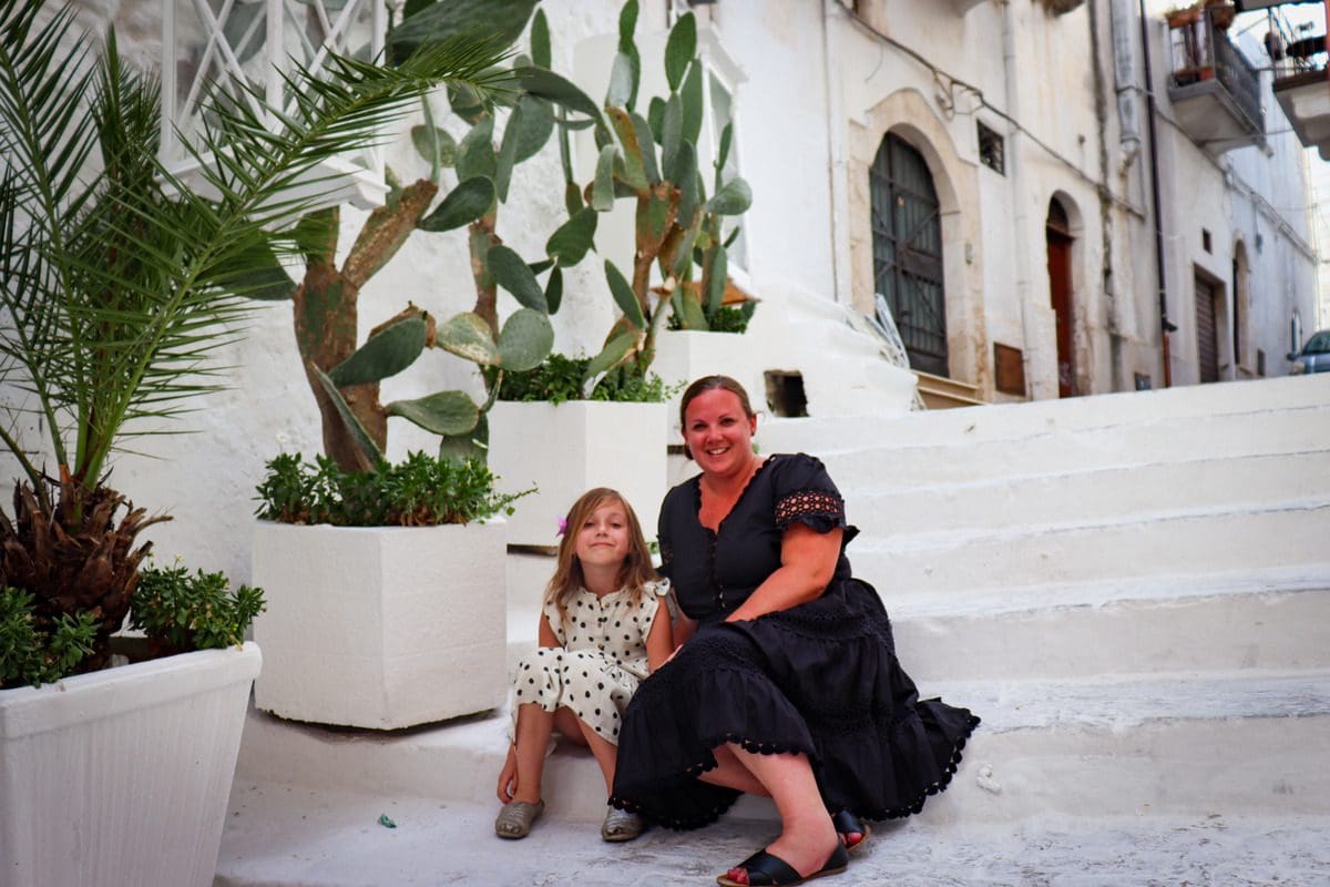 A mom and her young daughter sit together on white-washed steps in Ostuni.