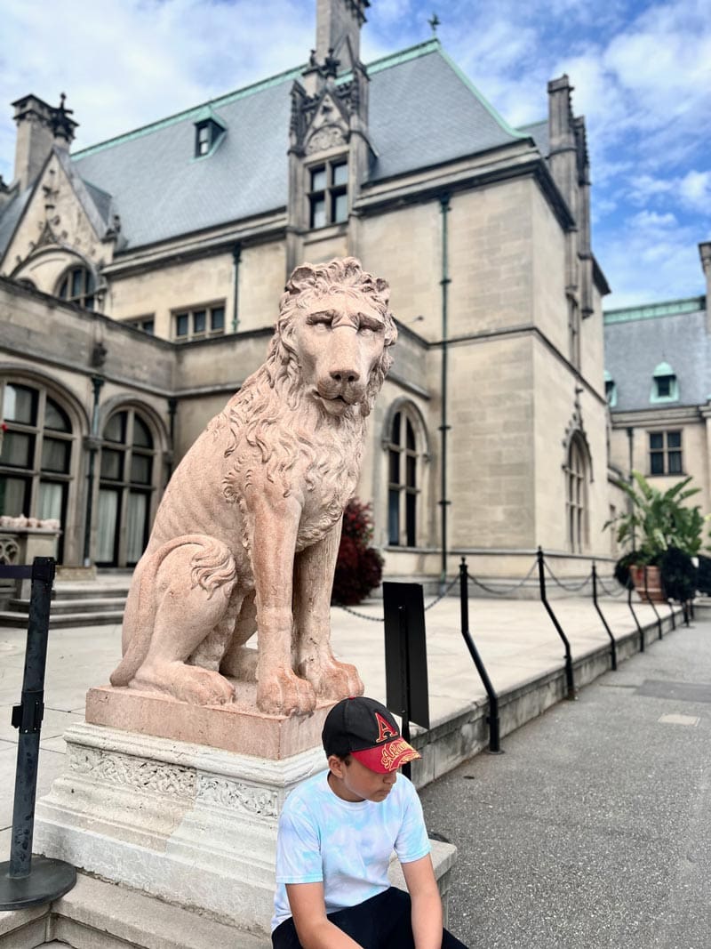 A young boy sits outside the Biltmore Estate near a lion statue, a must stop on any Asheville family vacation itinerary.