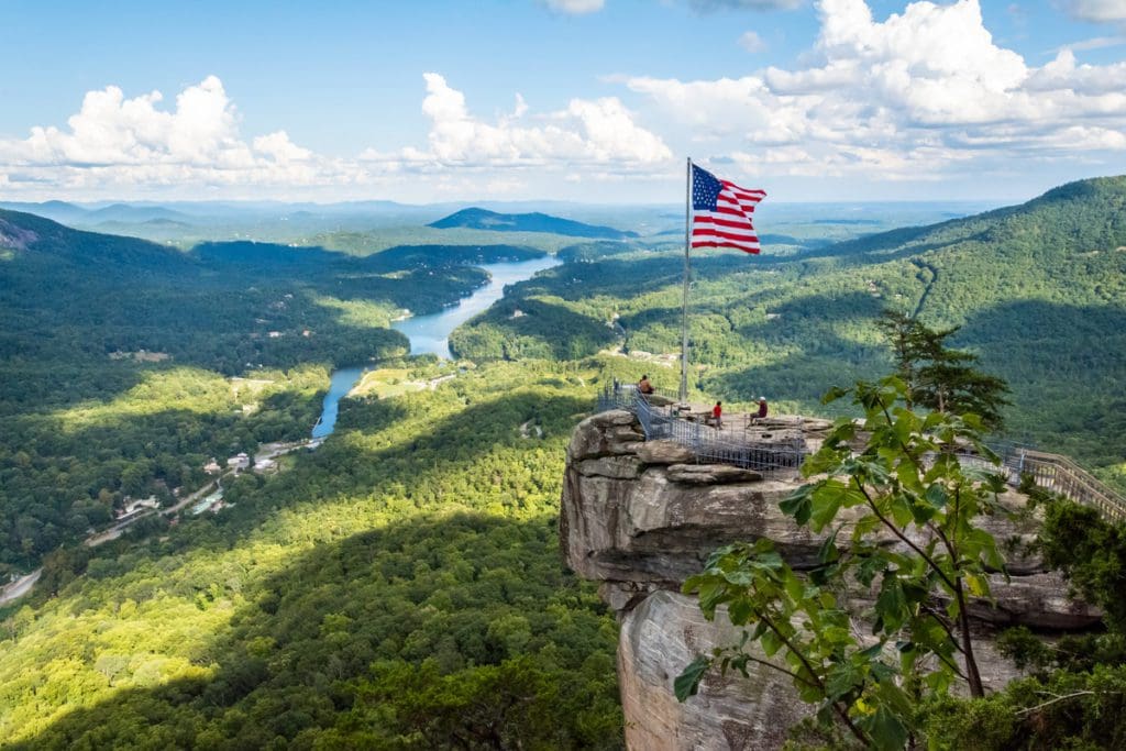 Several people walk around the top of Chimney Rock State Park on a sunny day, with a expansive view of the valley below.