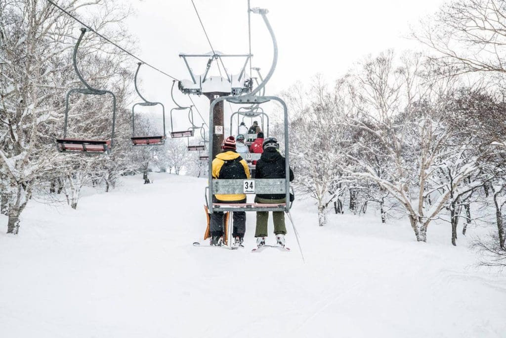 People sitting in a chair lift together going up the mountain on a winter's day at Nozawa Onsen Ski Resort .