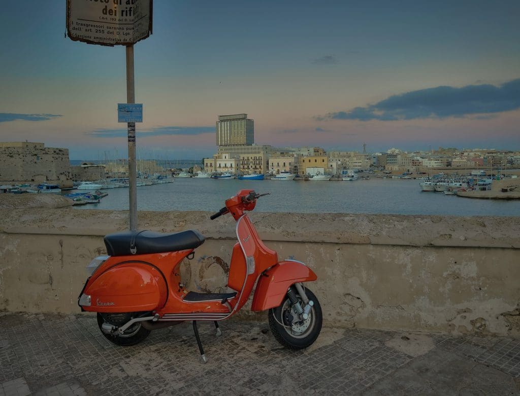 A red vespa parked along the Old Town wall of Gallipoli with the sea in the distance. 