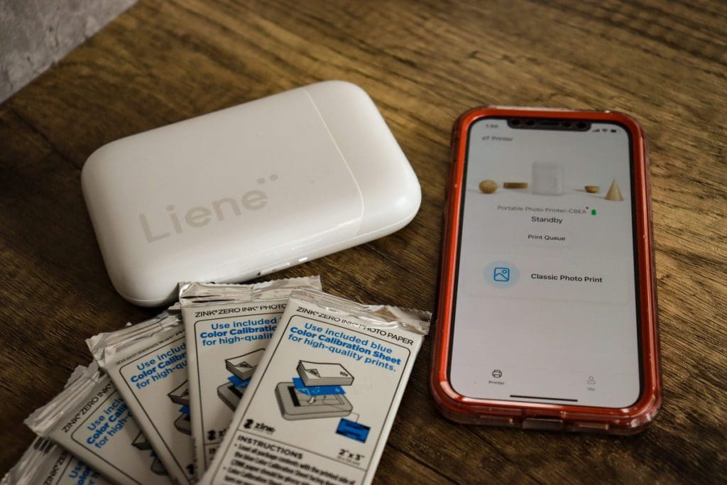 A product shot of a Liene Pearl Portable Photo Printer, with photo sheets near a iOS phone.