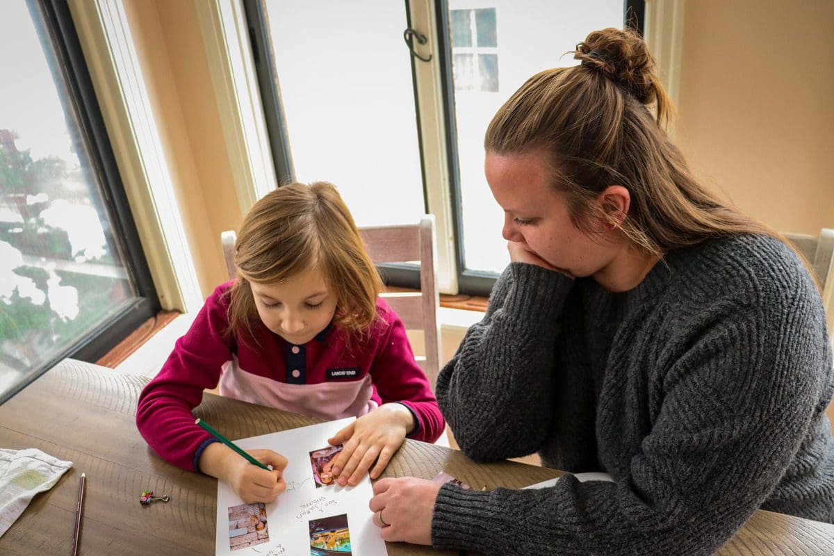 A mom watches on as her young daughter colors and creates a small travel diary.
