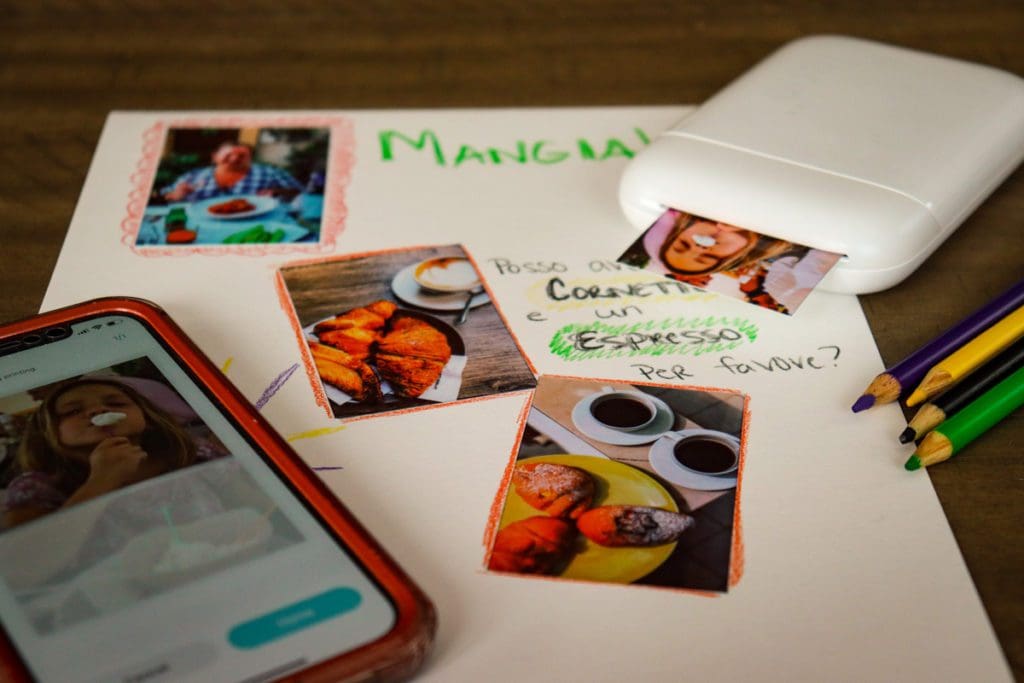 A Liene Pearl Portable Photo Printer on a table with a phone, printed images, and a sheet displaying a travel diary for food.