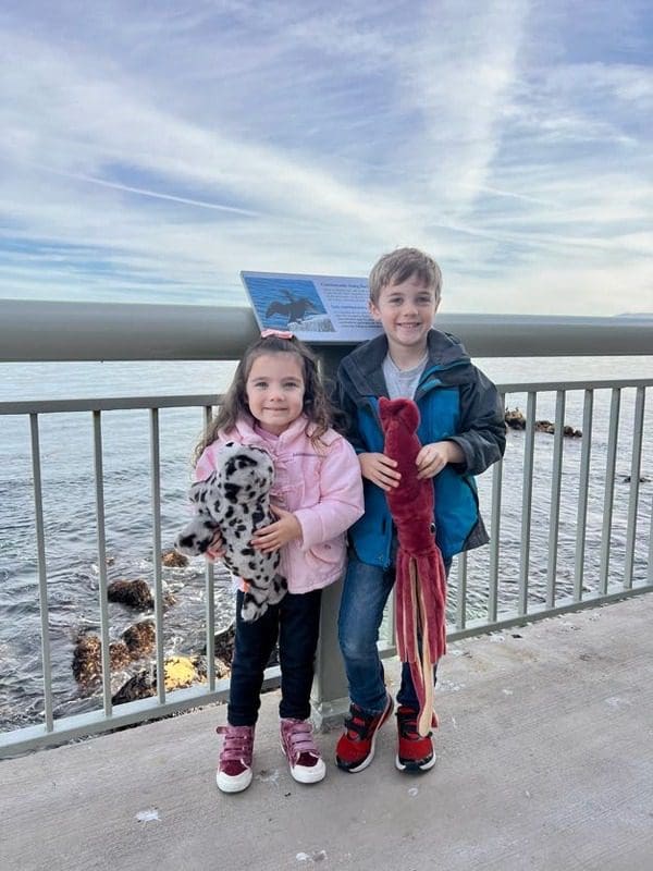 Two kids stand with new marine stuffies on the deck of Monterey Bay with the ocean in the distance.
