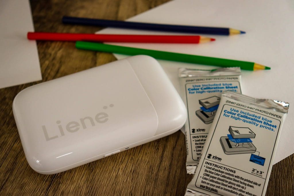 A product shot of the Liene Pearl Portable Photo Printer near the printer sheets, paper, and colored pencils.