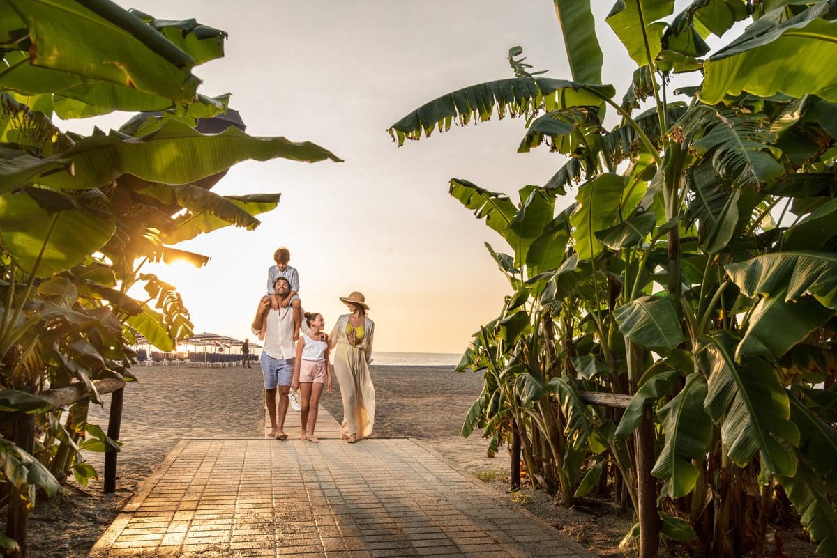 A family of four walks together across the beach at sunset while staying at Falkensteiner Club Funimation Garden Calabria.