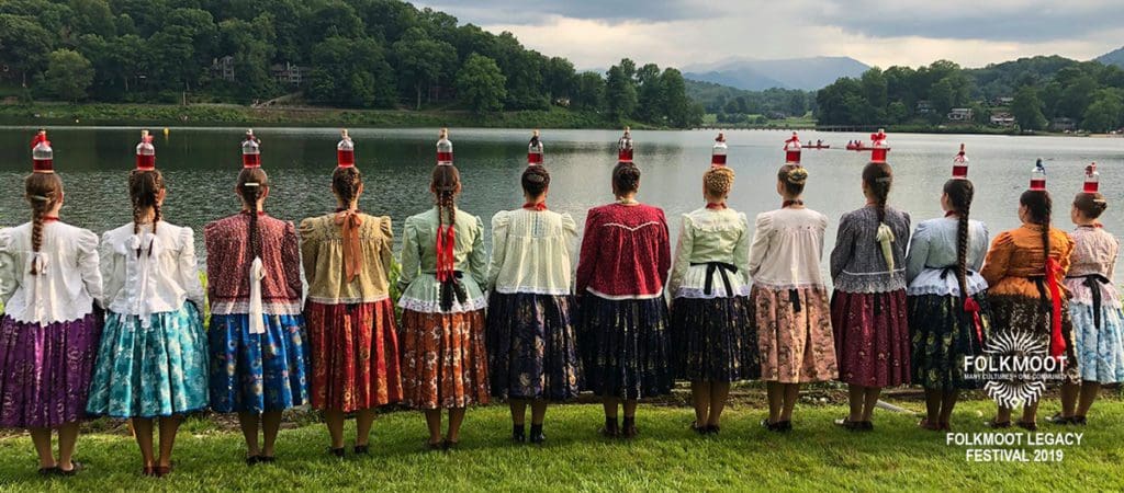Several women in traditional dance costumes stand together looking a lake near Waynesville, as part of Folkmoot USA.