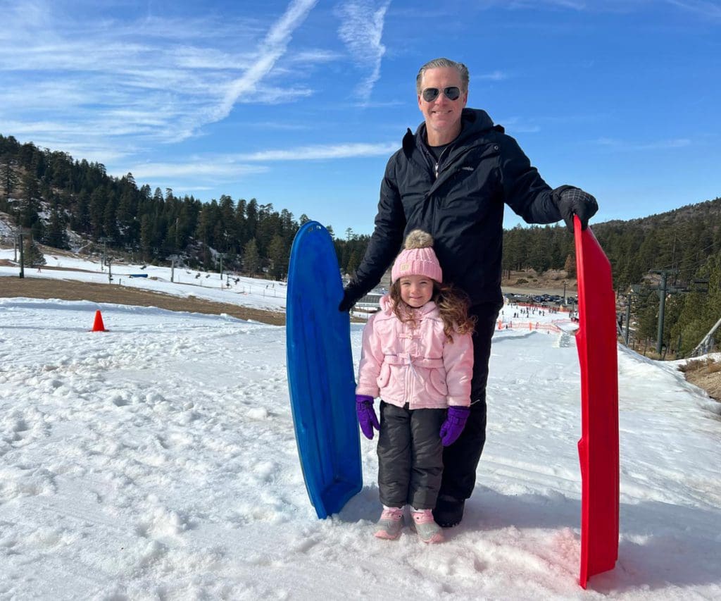A dad and his young daughter stand together in the snow with sleds.