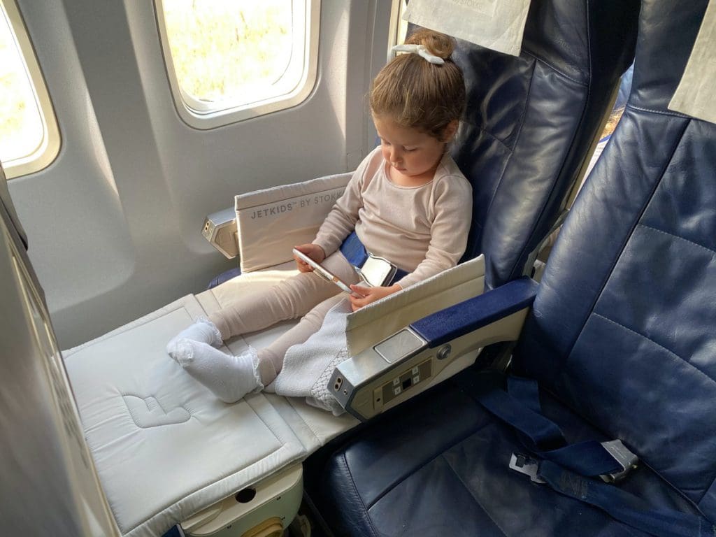 A young girl sits on a plan in her JetKids by Stokke, one of the best products for sleeping on long international flights with kids.