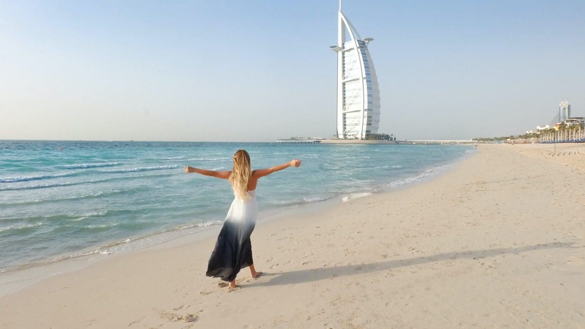 A woman gleefully throws up her arms while walking along the beach in Dubai, with the Burj Khalifa in the distance.