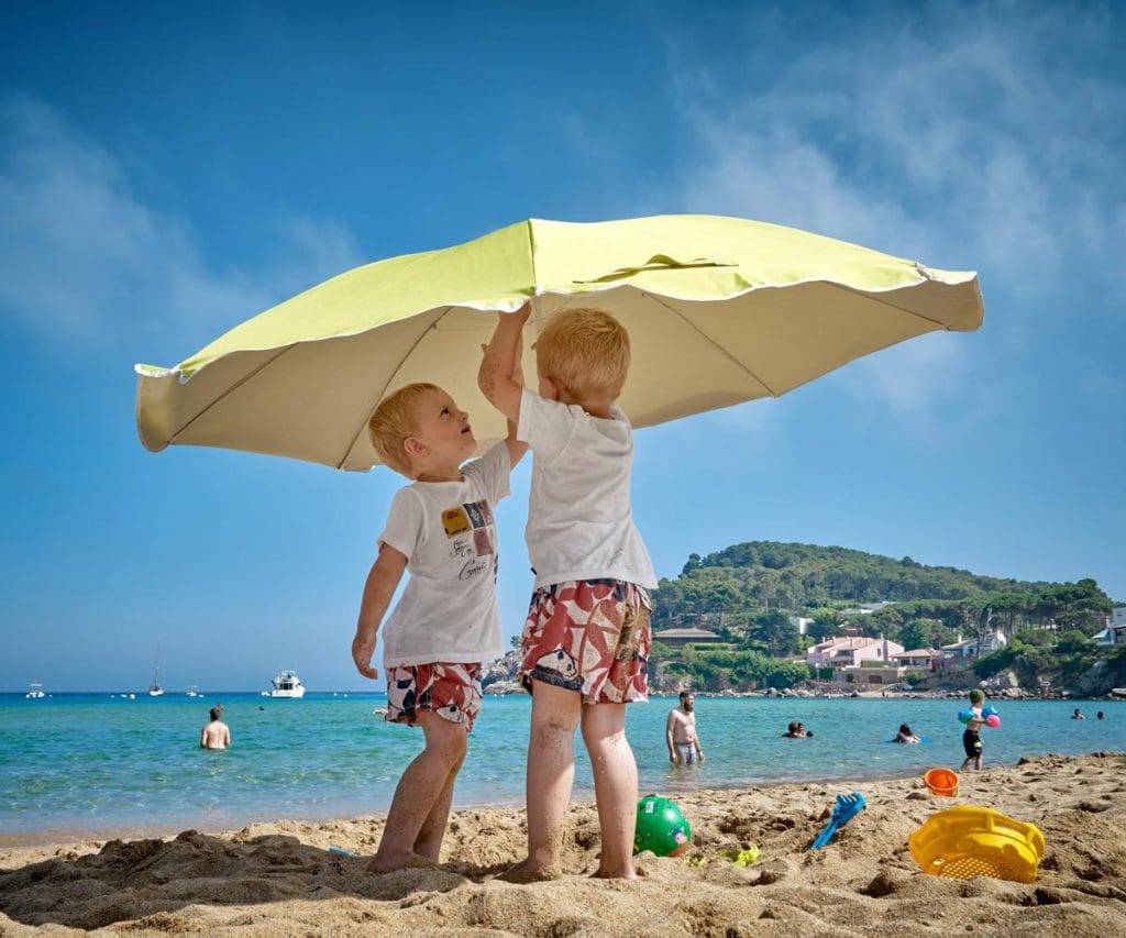 Two young boys play under a sun umbrella, while traveling with their family, a beach umbrella is a must on a beach packing list for families.
