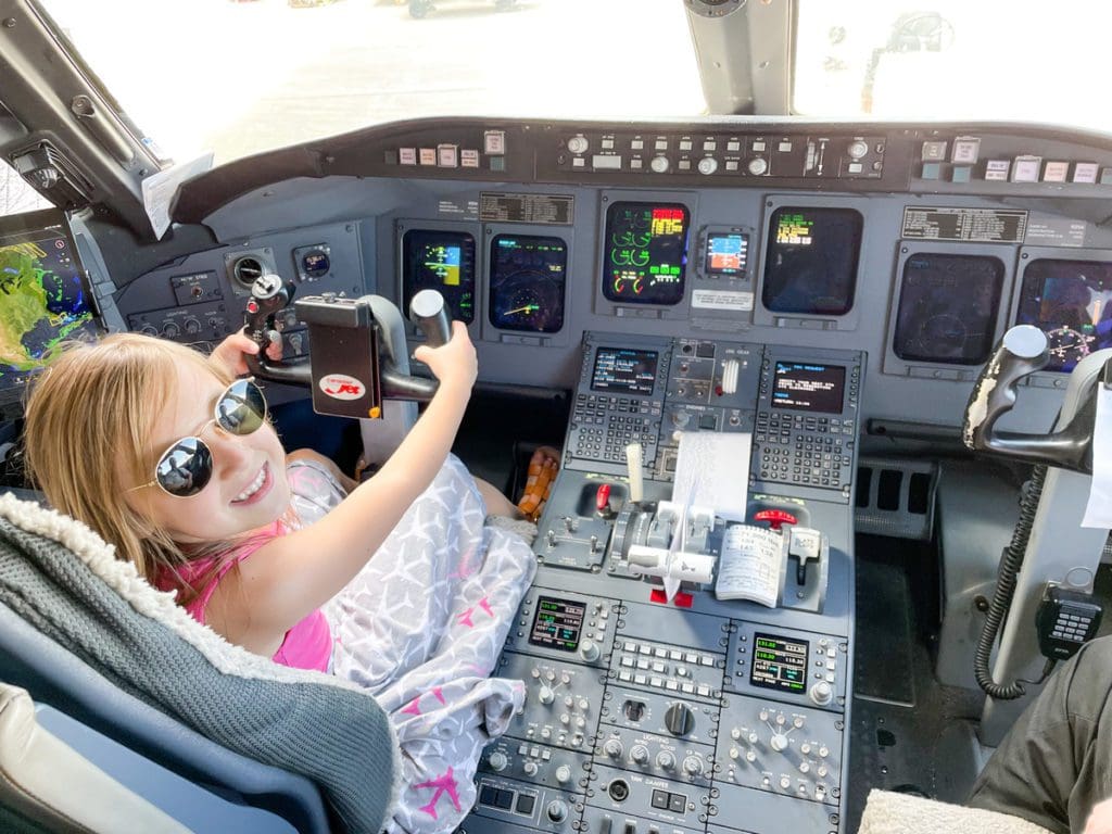 A young girl wearing aviators smiles back as she pretends to drive a Delta plane, while chatting with the plane's captain.
