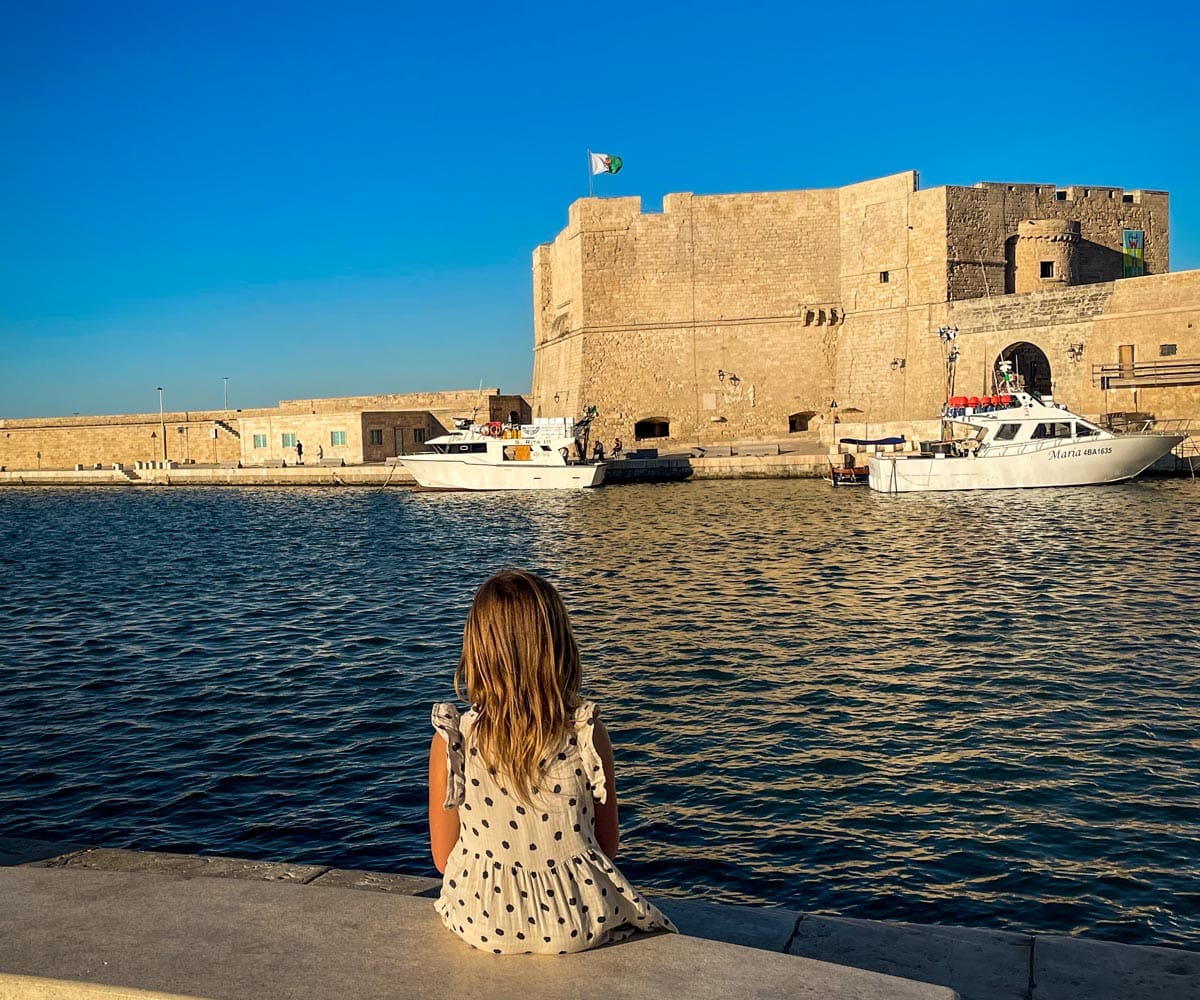 A young girl sits on a bench in the old sea port of Monopoli looking out onto the old city walls.