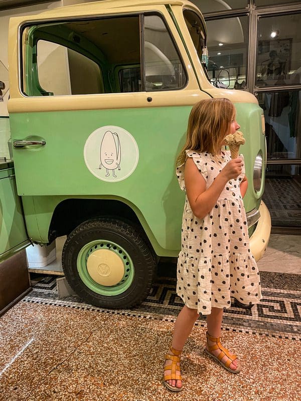 A young girl eats a gelato near a small green car.