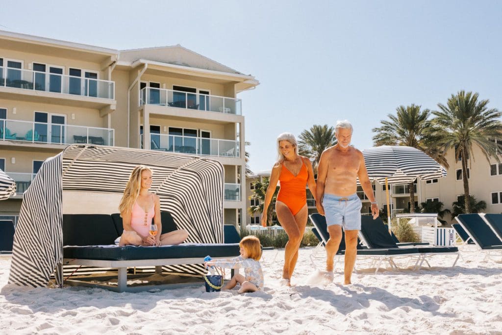 A mom and her young daughter sit on a beach cabana, while the grandparents stroll the beach nearby, at Edgewater Beach Hotel, one of the best hotels in Naples for families.