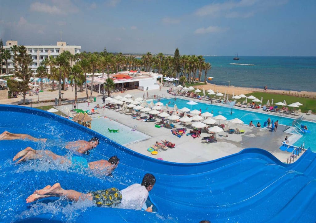 Several people race down a water slide at Louis Phaethon Beach, with resort buildings and grounds in the distance.