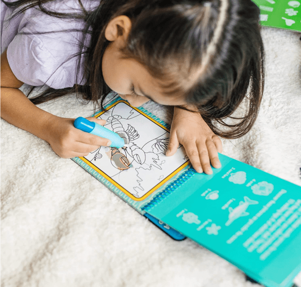 A young girl lays on the floor and colors in a Melissa & Doug Water Wow book, one of the best travel toys for young kids.