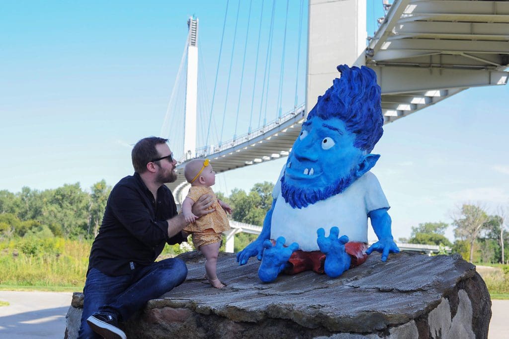 An infant and a dad look at the blue troll statue under the pedestrian bridge in Omaha, one of the best affordable summer vacations in the United States with kids.