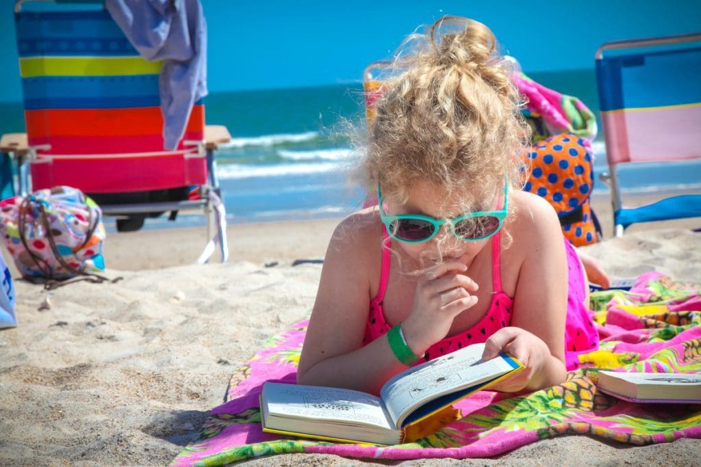 A young girl wearing sunglasses, lies on a beach towel reading a book on a sunny day at the beach with the ocean in the distance.