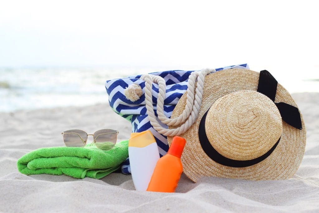 A beach bag with a hat, sunscreen, towel, and sunglasses rest on a sandy beach, which are necessary on any beach packing list for families.