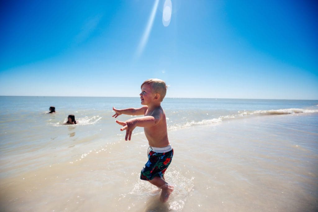 A happy swimming toddler reaching his hands out toward the ocean on Sanibel Beach, one of the best Florida beaches for families.
