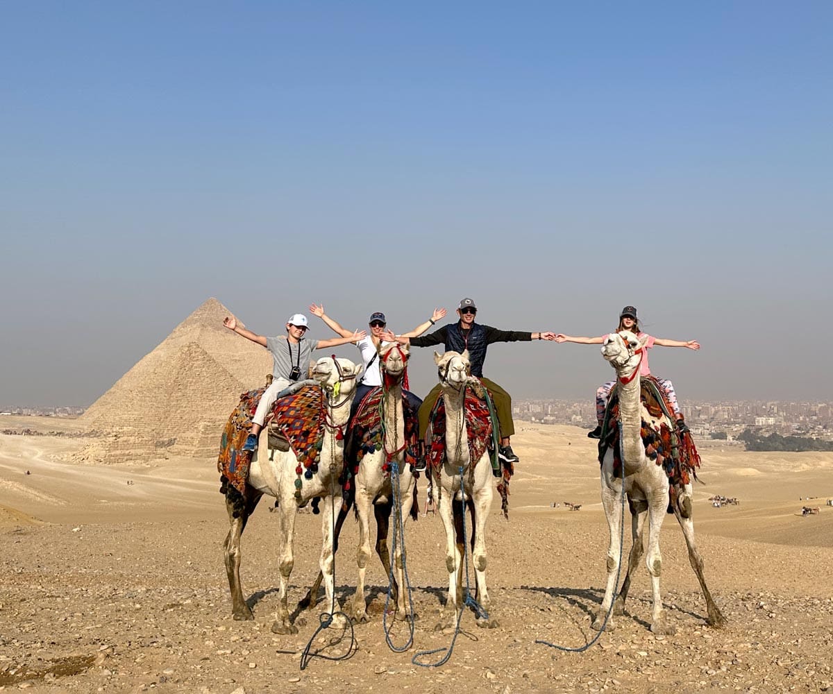 A family of four sits atop camels in front of the Pyramids of Giza.
