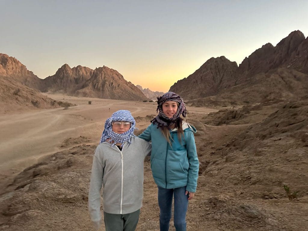 Two kids stand together, both wearing head scarves, on a desert tour in Egypt, a must on this 2-Week Egypt Itinerary for Families.