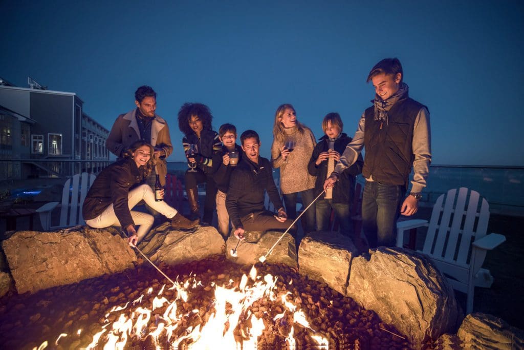 A large group of guests roasts marshmallows in an on-site fire pit at Cliff House Maine.