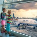 Two kids in brightly colored clothing stand together looking out of a airport window at a United Airlines plane.