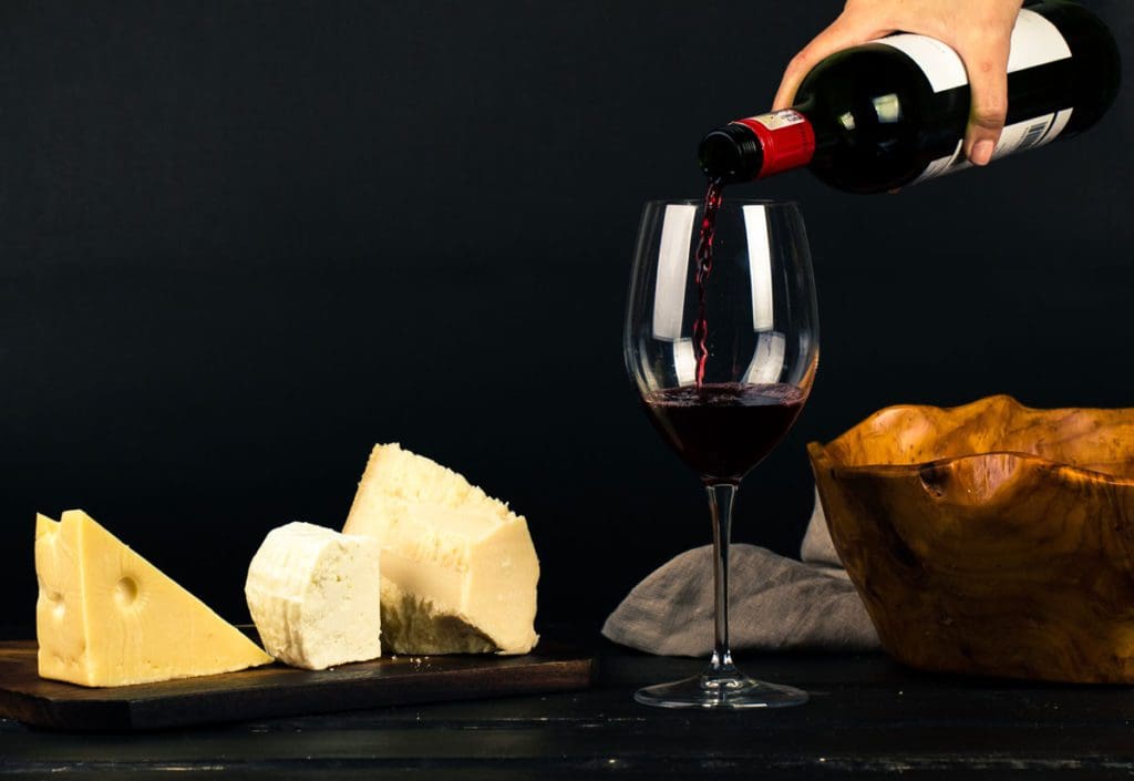 A hand pours a glass of red whine on a table filled with different types of cheese.