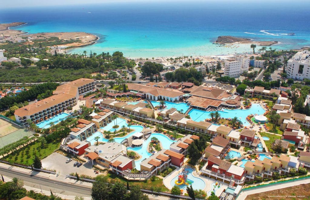 An aerial view of Atlantica Aeneas Resort, featuring several pools, lush grounds, and buildings, with the ocean in the distance.