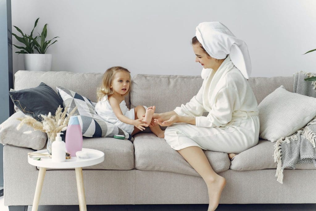 A mom and her young daughter sit on a couch together enjoy an at-home spa day.