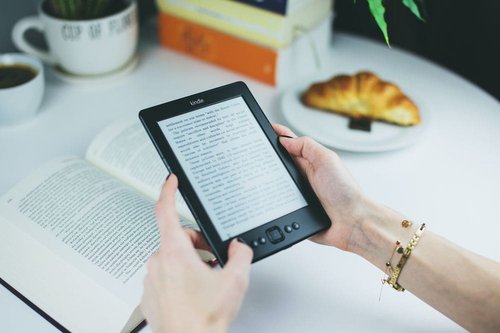 Two hands reach out to hold a Kindle over a table, near a cup of coffee and a croissant.