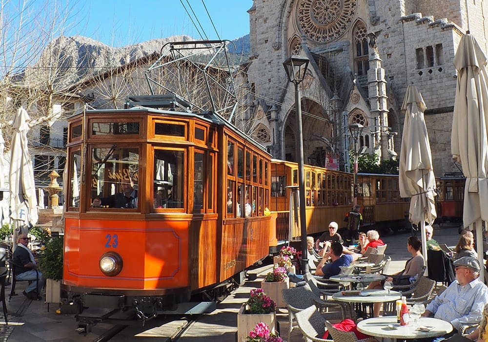 Tren de Sóller moves through the city near a cafe crowded with people.