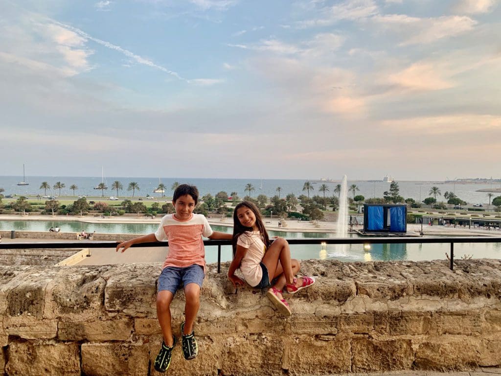 Two kids sit along a stone wall with a view of Palma behind them.