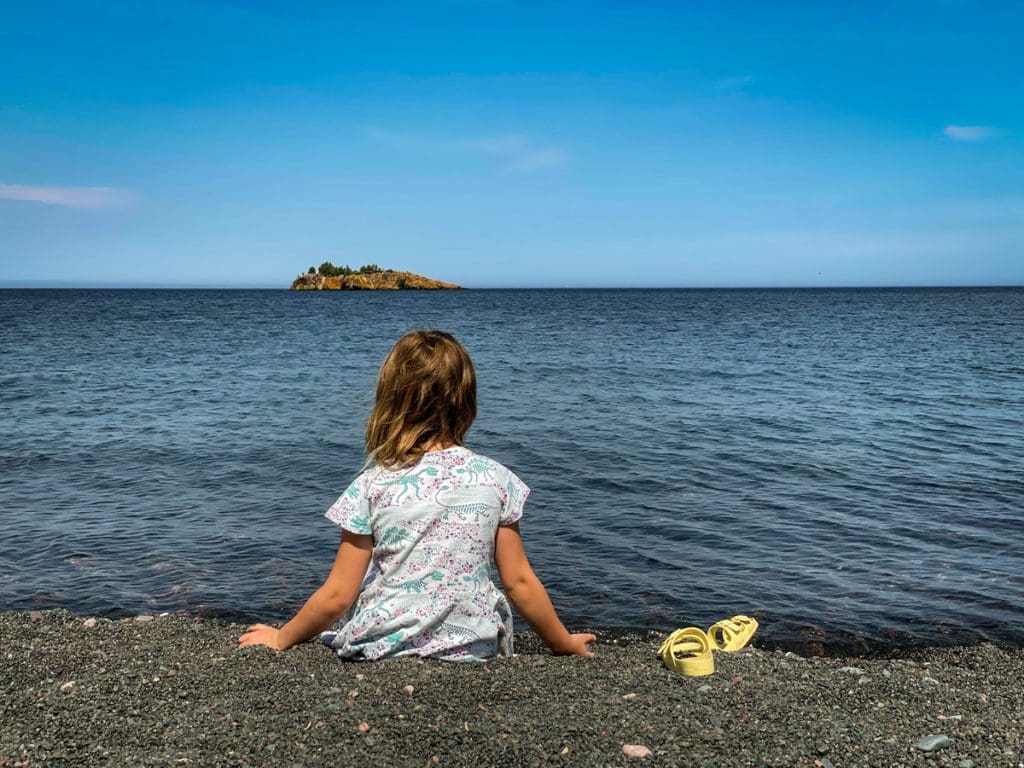 A young girl sits on a black pebble beach looking out toward the water.