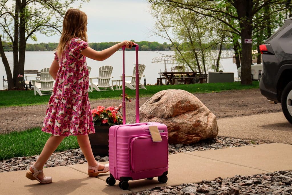 A young girl walks along a sidewalk toward a car with pink luggage.