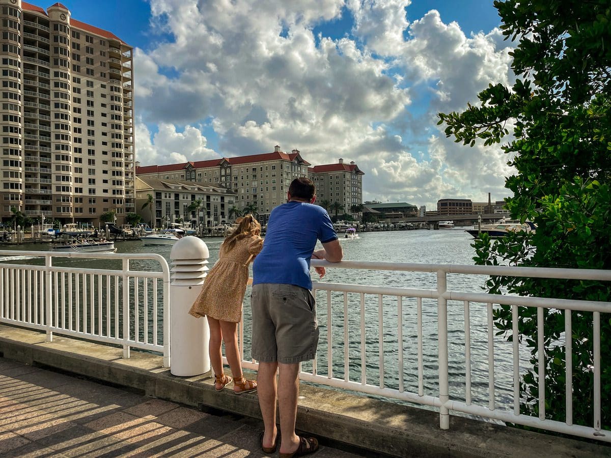 A dad and his young daughter lean over a railing to look at the river, while strolling along the Tampa Riverwalk.
