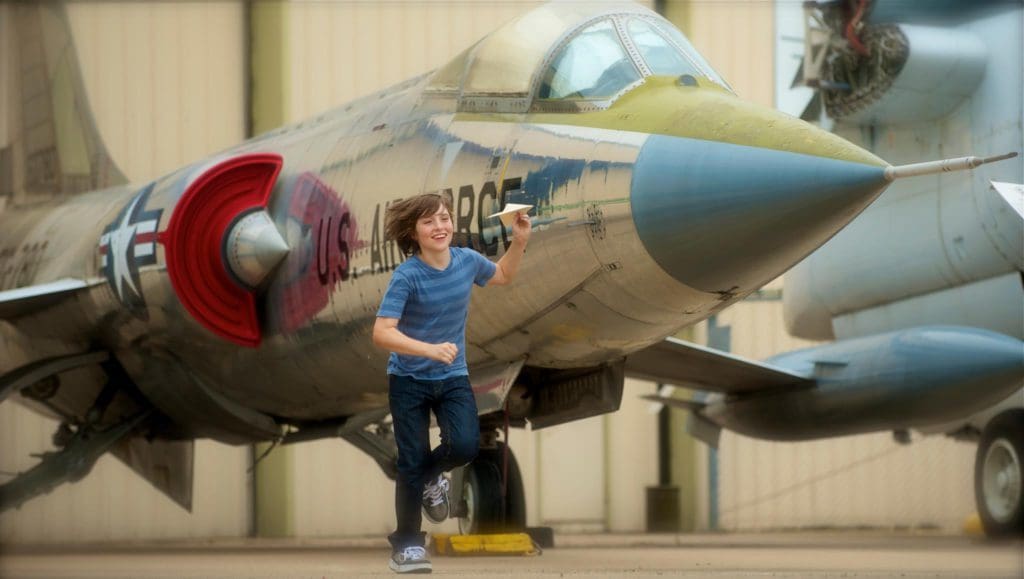 A young boy runs with a paper airplane alongside a military plane at Cavanaugh Flight Museum.