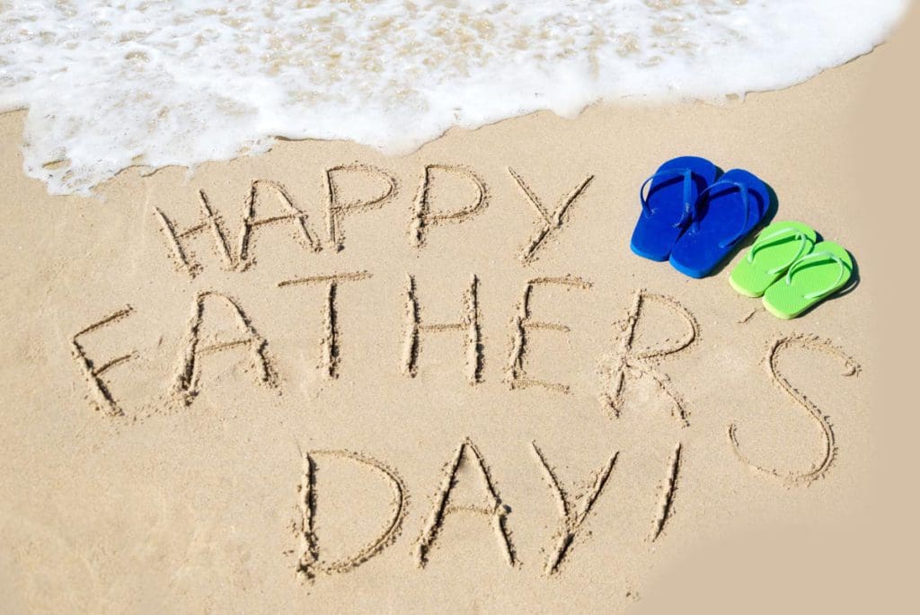 "Happy Father's Day" written in the sand with sandals nearby.