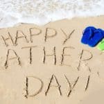 "Happy Father's Day" written in the sand with sandals nearby.