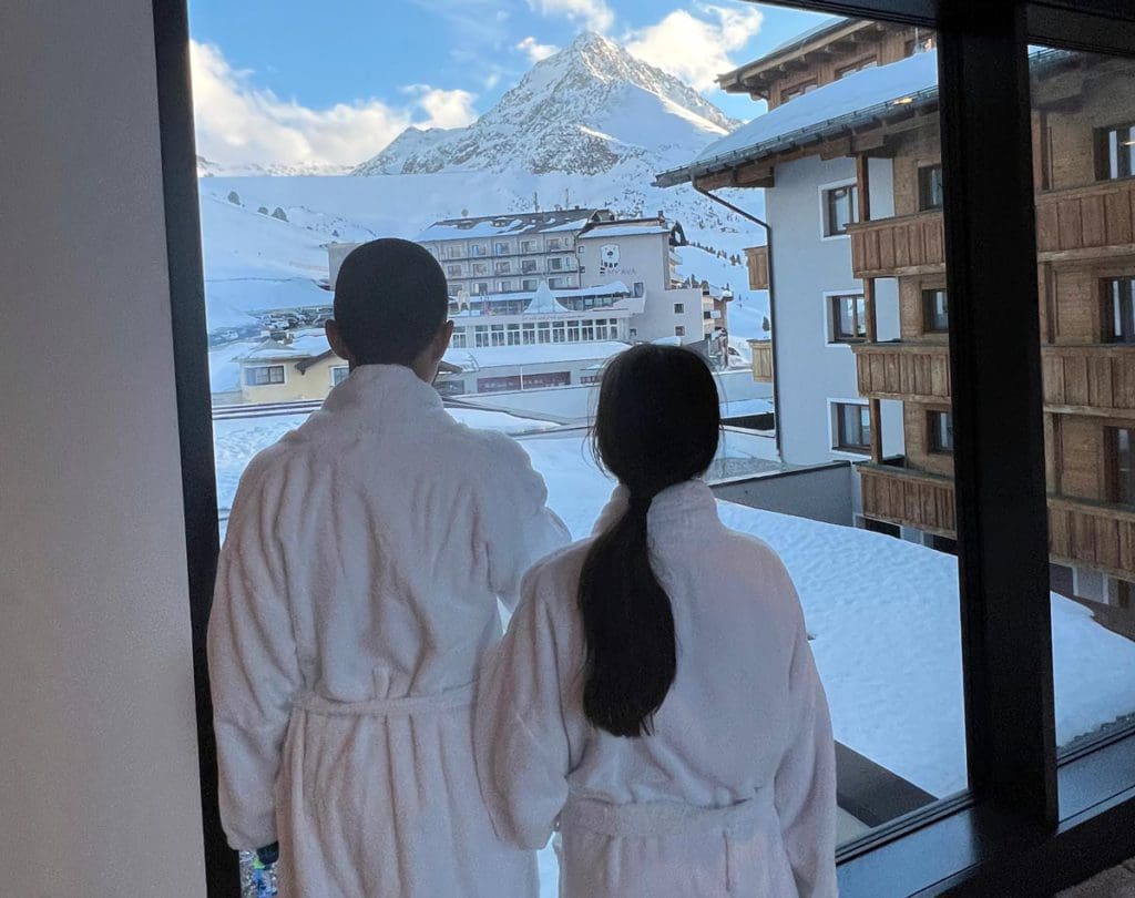 Two kids in bathrobes stand at a window looking out onto a mountain scenery in the Austrian Alps.