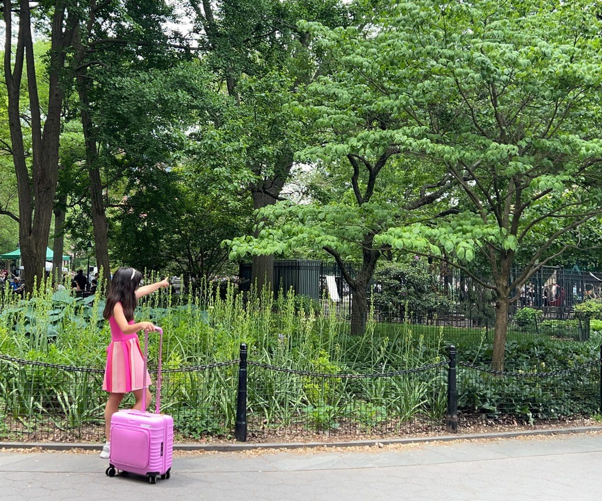 A young girl walks through a park with her suitcase, pointing to something far off.